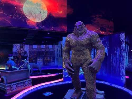 A large, furry Bigfoot statue stands in a colorful, illuminated exhibit with a dramatic moonlit backdrop.