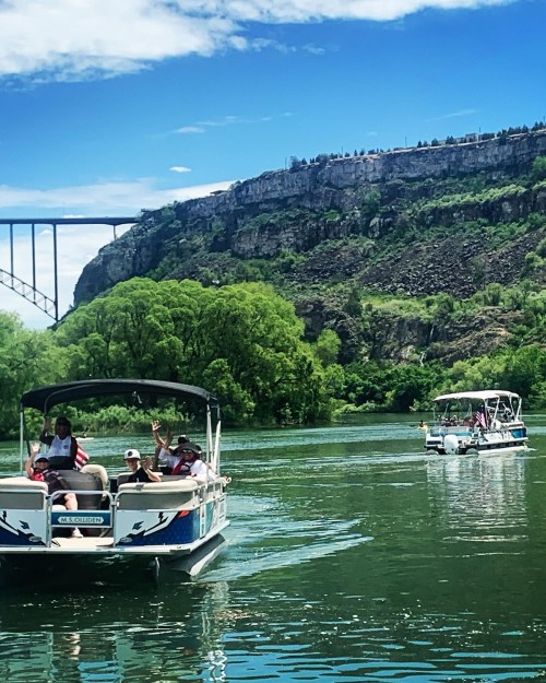 Two boats on a river, surrounded by lush greenery and a rocky cliff under a blue sky with scattered clouds.