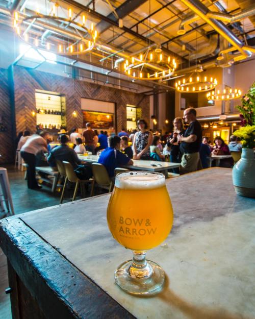 A glass of beer sits on a table in a lively brewery with people socializing and warm lighting in the background.