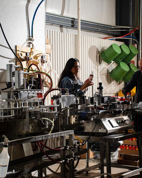 A woman holds a phone in a factory setting, with machinery and green containers in the background.