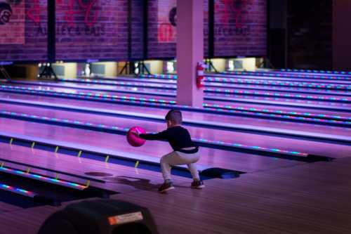 A child prepares to bowl, holding a pink bowling ball in a brightly lit bowling alley.