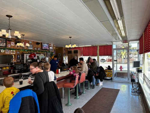 A busy diner interior with families seated at the counter and booths, enjoying meals and drinks. Bright, cheerful atmosphere.