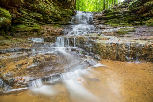 A serene waterfall cascades over rocky terrain, surrounded by lush green foliage in a tranquil forest setting.