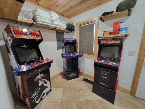 Three arcade machines in a cozy room: NBA Jam, a multi-game console, and Mortal Kombat, with towels stacked nearby.