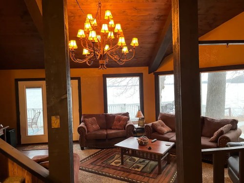 Cozy living room with a chandelier, brown sofas, and a view of a lake through large windows. Warm, inviting atmosphere.