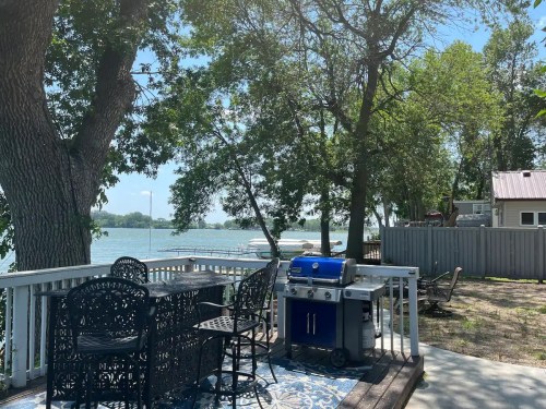 A lakeside patio with a grill, dining table, and chairs, surrounded by trees and a view of the water.