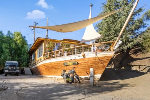 A unique wooden house resembling a boat, with a sail, surrounded by trees and a vintage motorcycle parked nearby.