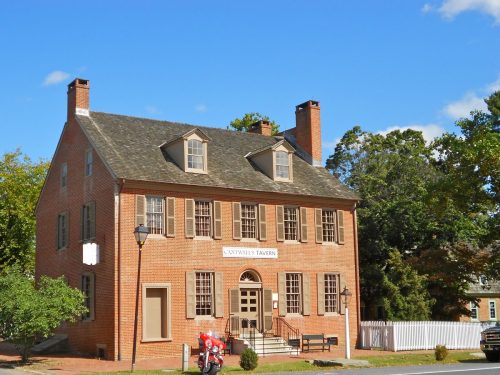 Historic brick building with a gabled roof, labeled "Gadsby's Tavern," surrounded by trees and a white picket fence.