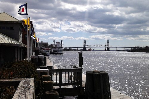 A scenic waterfront view with boats, a bridge, and cloudy skies, featuring a wooden dock and colorful flags.