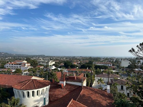 A panoramic view of a coastal city with red-tiled roofs, palm trees, and a blue sky with wispy clouds.
