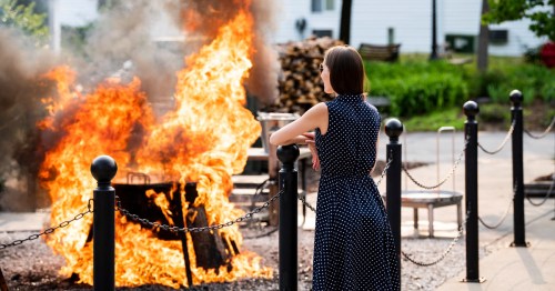 A woman in a polka dot dress stands near a fire, observing the flames and smoke in a park setting.