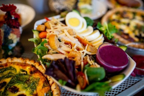 A colorful salad with sliced eggs, chicken, cheese, and a purple dressing, surrounded by various quiches and dishes.