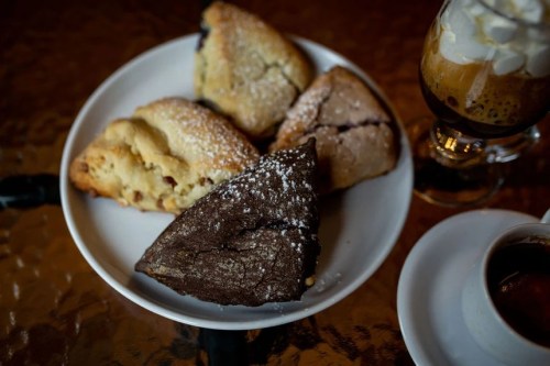 A plate of assorted pastries, including scones and a chocolate treat, next to a glass of coffee.