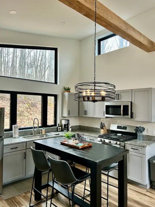 Modern kitchen with gray cabinets, a black island, and large windows overlooking a wooded area. Stylish pendant light above.