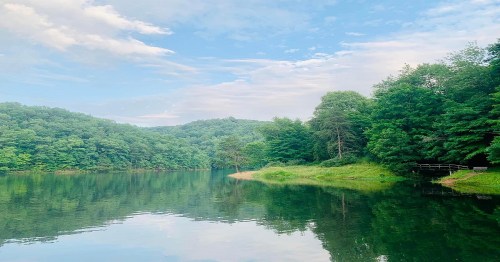 A serene lake surrounded by lush green trees under a blue sky with soft clouds, reflecting the landscape in the water.