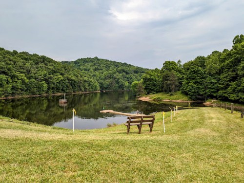A serene lake surrounded by lush green trees, with a wooden bench overlooking the water and a small dock in the distance.