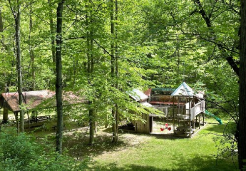A wooden cabin surrounded by lush green trees, featuring a deck and a small play area with a slide.