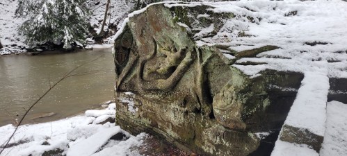 A snow-covered rock with intricate carvings beside a flowing river in a wooded area.