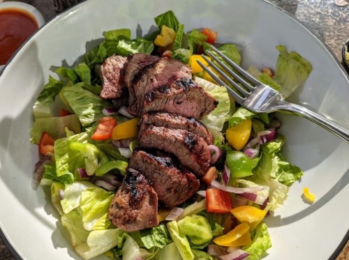 A bowl of fresh salad topped with sliced grilled steak, featuring lettuce, tomatoes, and bell peppers.