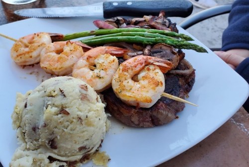 A plate with grilled shrimp, asparagus, a steak, and mashed potatoes, garnished with a knife in the background.