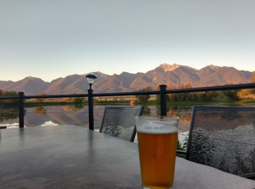 A glass of beer on a table with mountains and a calm lake in the background during sunset.