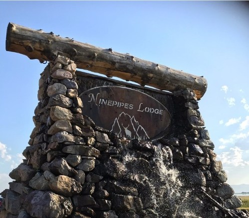 Sign for Ninepipes Lodge made of logs and stones, with a water feature in front, set against a clear blue sky.