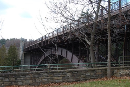 A large metal bridge arches over a river, surrounded by trees and a stone wall, under a cloudy sky.