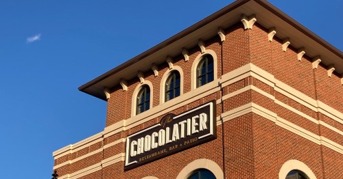 A brick building with a sign reading "The Chocolatier Restaurant, Bar & Patio" against a clear blue sky.