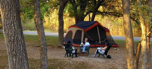 A campsite with a large tent and two people sitting around a fire pit, surrounded by trees and autumn foliage.
