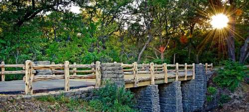 A wooden bridge spans a stone structure, surrounded by lush greenery and illuminated by a bright sunset.