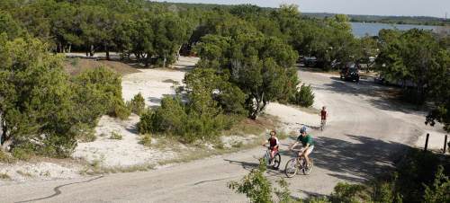 Two cyclists ride along a winding dirt path surrounded by trees, with a lake visible in the background.
