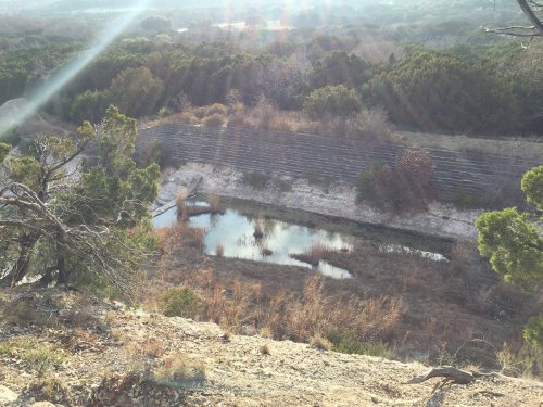 A scenic view of a dry landscape with a small pond surrounded by trees and brush, under a bright sky.