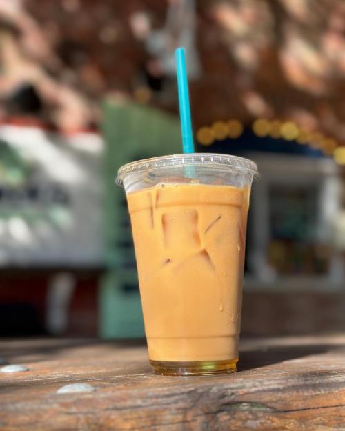 A clear cup of iced coffee with a blue straw sits on a wooden table, with a blurred background.