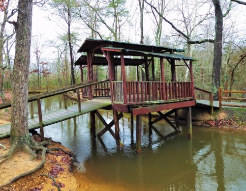A wooden gazebo on stilts over a calm pond, surrounded by bare trees and a dirt path.