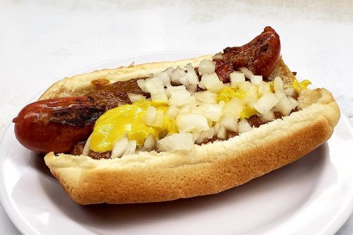 A hot dog in a bun topped with chili, mustard, and chopped onions on a white plate.