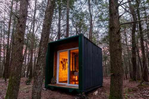 A small black cabin nestled among tall trees, with warm light glowing from its interior at dusk.