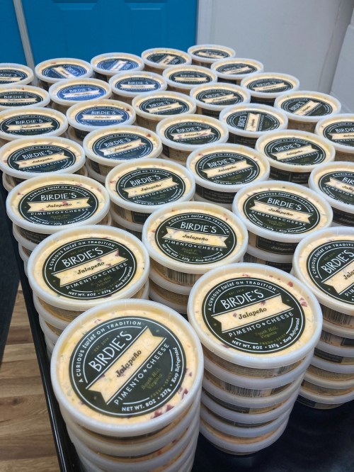 A table stacked with containers of Birdie's jalapeño pimento cheese, featuring black labels and a colorful design.