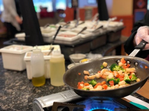 A chef stirs a colorful vegetable and shrimp stir-fry in a pan over a flame, with a buffet in the background.