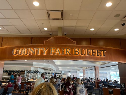 Sign for "County Fair Buffet" in a spacious dining area with people seated and a buffet setup in the background.