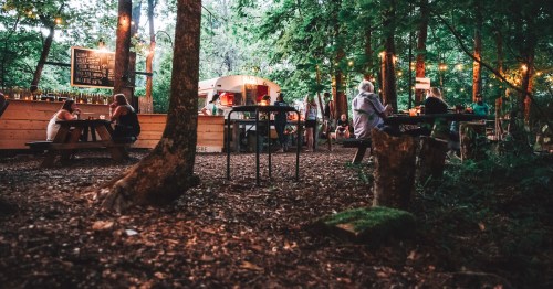 A cozy outdoor setting in a forest with string lights, picnic tables, and a food truck, filled with people enjoying the atmosphere.