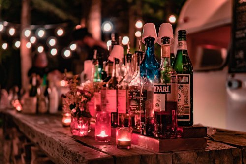 A rustic bar with various liquor bottles and candles, illuminated by string lights in a cozy outdoor setting.
