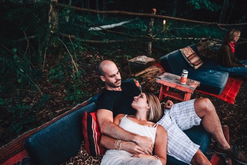 A couple relaxes together on a couch in a forest setting, with a woman gazing at the man, while another person sits nearby.