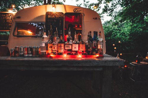 A rustic bar setup with various bottles and cans, illuminated by candles, in front of a vintage trailer at dusk.
