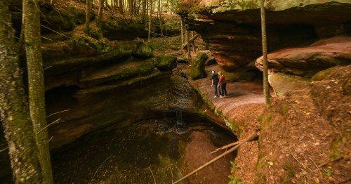 Two people stand by a serene, wooded area with rock formations and a small pond. Sunlight filters through the trees.