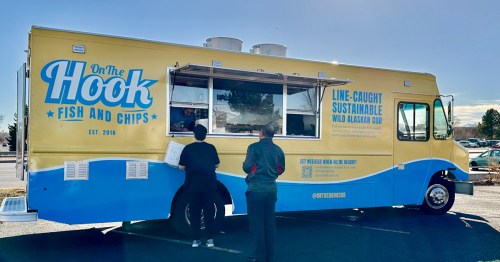 A bright blue and yellow food truck named "On the Hook" serves fish and chips, with customers ordering at the window.