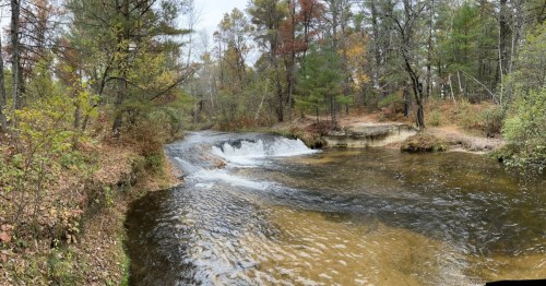 A serene river flows through a forested area, with gentle waterfalls and autumn foliage surrounding the scene.