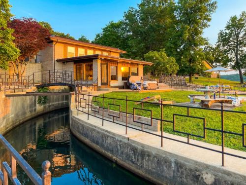 A modern house by a calm waterway, surrounded by greenery and outdoor seating, under a clear blue sky.