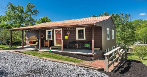 A cozy brown house with a metal roof, porch seating, and landscaped yard under a clear blue sky.