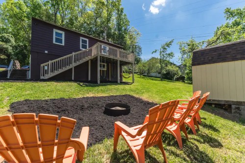 A backyard scene featuring orange chairs around a fire pit, with a house and trees in the background under a blue sky.