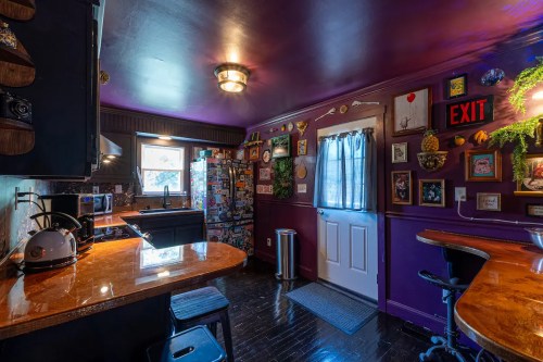 A cozy kitchen with purple walls, wooden accents, and eclectic decor, featuring a window, door, and bar seating.